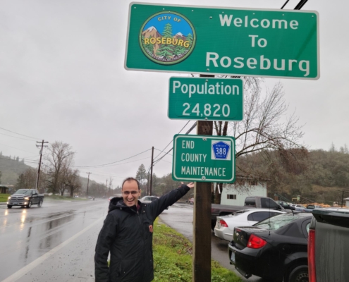 Sami stands by a sign reading Welcome to Roseburg, Population 24,820