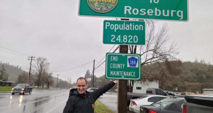 Sami stands by a sign reading Welcome to Roseburg, Population 24,820